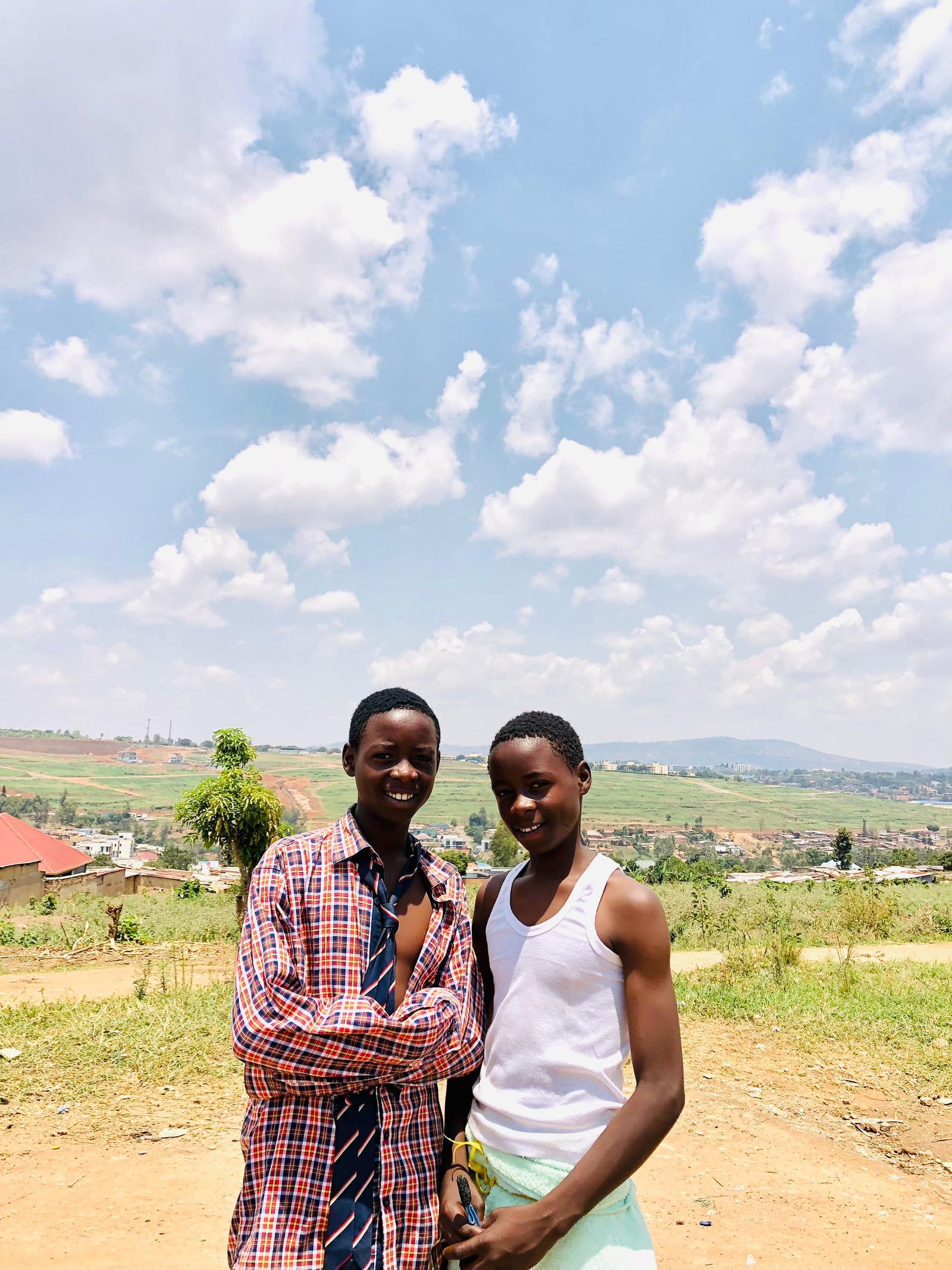Two smiling young dancers on a red-earth road with the green hills of Kigali behind them.